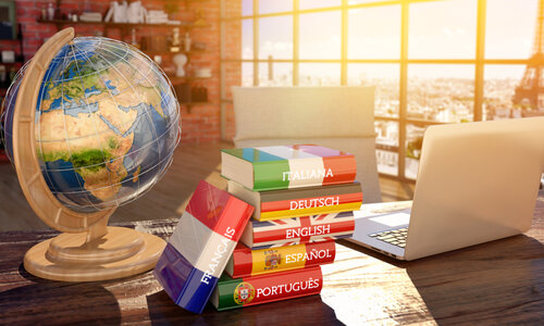 Die Sprachen auf dem Schreibtisch Auf einem Holzschreibtisch stehen ein Laptop, eine Weltkugel und mehrere Wörterbücher großer europäischer Sprachen (Italienisch, Deutsch, Englisch, Spanisch, Portugiesisch und Französisch). Durch die Fensterfront scheint intensives Sonnenlicht in den Raum und hüllt den Schreibtisch in ein fast märchenhaftes Licht.