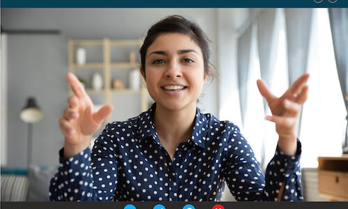 Im Web-Meeting Eine junge Frau in einer dunkelblauen Bluse mit weißen Punkten sitzt vor einem Bildschirm und gestikuliert mit den Armen. Sie scheint Teilnehmerin eines Web-Meetings zu sein. Im Hintergrund ist ein minimalistisch eingerichtetes Zimmer erkennbar.