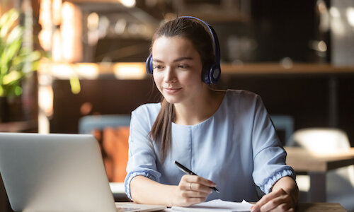 Frau am Schreibtisch Eine junge Frau in einem schicken, hellblauen Oberteil sitzt am Schreibtisch. Sie trägt ein Headset und schaut aufmerksam in den Laptop, der vor ihr steht. Sie hat einen Stift in der Hand und ein Stück Papier liegt auf dem Tisch. Am Fenster im Hintergrund steht eine Grünpflanze, die man nur verschwommen erkennen kann.