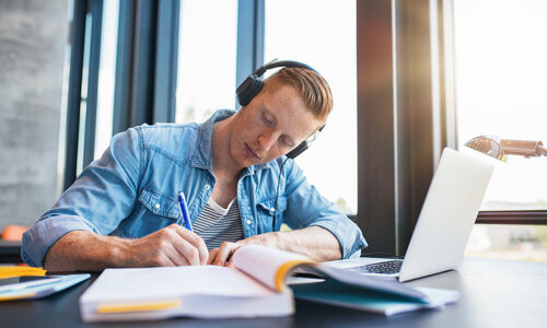 Student beim Lernen Ein junger Student sitzt an einem Schreibtisch direkt am Fenster und macht Notizen. Auf dem Tisch befinden sich ein Laptop und mehrere Bücher. Der Mann trägt ein lässiges Jeanshemd, darunter ein geringeltes Shirt. Er hat große, schwarze Kopfhörer auf.