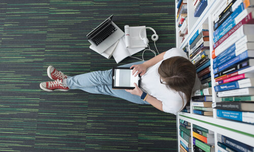 Person sitzt mit Tablet auf dem Boden vor einem Bücherregal, umgeben von Laptop, Papieren und einem Poster