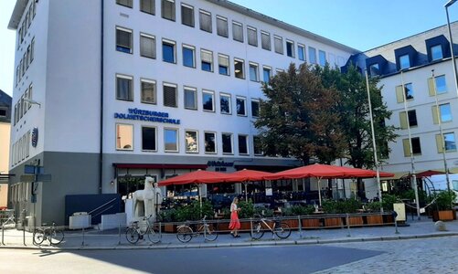 Ein mehrstöckiges Gebäude mit zahlreichen Fenster. Im Erdgeschoss ein Café mit Außenbereich und Sonnenschirmen.