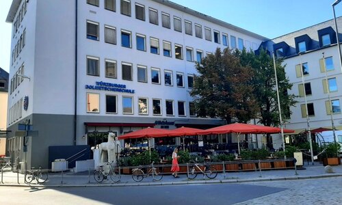 Die Würzburger Dolmetscherschule von außen Ein mehrstöckiges Gebäude mit zahlreichen Fenster. Im Erdgeschoss ein Café mit Außenbereich und Sonnenschirmen.