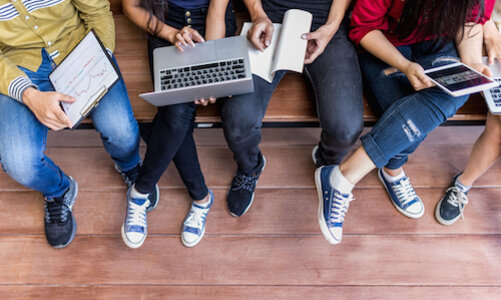 Blick aus der Vogelperspektive auf eine Gruppe von Schüler*innen. Sie tragen Jeans und Sneaker, in ihren Händen (über den Knien) halten sie Laptops, ein Tablet, Bücher. Die Gesichter sind außerhalb des Bildes, man kann aber bei den Frauen lange Haare erkennen.