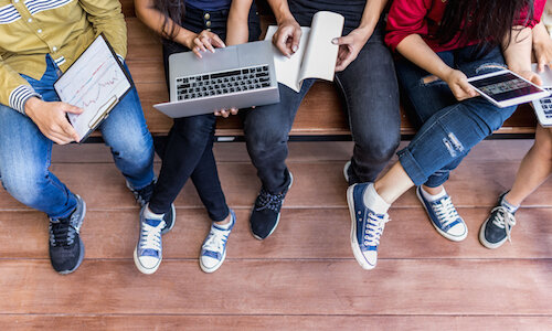 Schüler*innen von oben Blick aus der Vogelperspektive auf eine Gruppe von Schüler*innen. Sie tragen Jeans und Sneaker, in ihren Händen (über den Knien) halten sie Laptops, ein Tablet, Bücher. Die Gesichter sind außerhalb des Bildes, man kann aber bei den Frauen lange Haare erkennen.