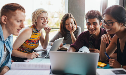 Fünf Personen sitzen an einem Tisch und arbeiten gemeinsam mit Laptops, Notizbüchern und Papieren