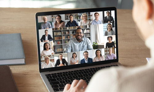 Online-Meeting An der Schulter und Gesicht einer Person kann man auf deren Laptopbildschirm blicken. Dort tummeln sich fünfzehn Gesichter, eins davon groß in der Mitte. Die Szene zeigt ein Online-Meeting.