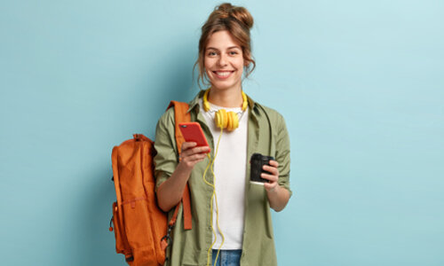 Frau mit Handy und Kaffeebecher Eine Frau steht vor einer türkisen Wand. Sie hat einen orangefarbenen Rucksack umhängen, um den Hals hängen Kopfhörer, in ihrer rechten Hand hält sie ein orangefarbenes Handy, links einen Kaffeebecher.