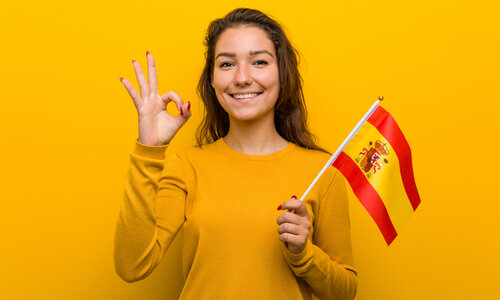 Spanien Ein junges Mädchen mit langen Haaren steht in einem gelben Pullover vor einer gelben Wand, ihre Fingernägel sind rot lackiert. In ihrer linken Hand hält sie die gelb-rote spanische Flagge, mit der rechten Hand signalisiert sie das "Perfekt"-Zeichen (Daumen und Zeigefinger sind zu einem Kreis geformt, die anderen Finger nach oben gereckt).