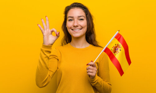 Ein junges Mädchen mit langen Haaren steht in einem gelben Pullover vor einer gelben Wand, ihre Fingernägel sind rot lackiert. In ihrer linken Hand hält sie die gelb-rote spanische Flagge, mit der rechten Hand signalisiert sie das "Perfekt"-Zeichen (Daumen und Zeigefinger sind zu einem Kreis geformt, die anderen Finger nach oben gereckt).