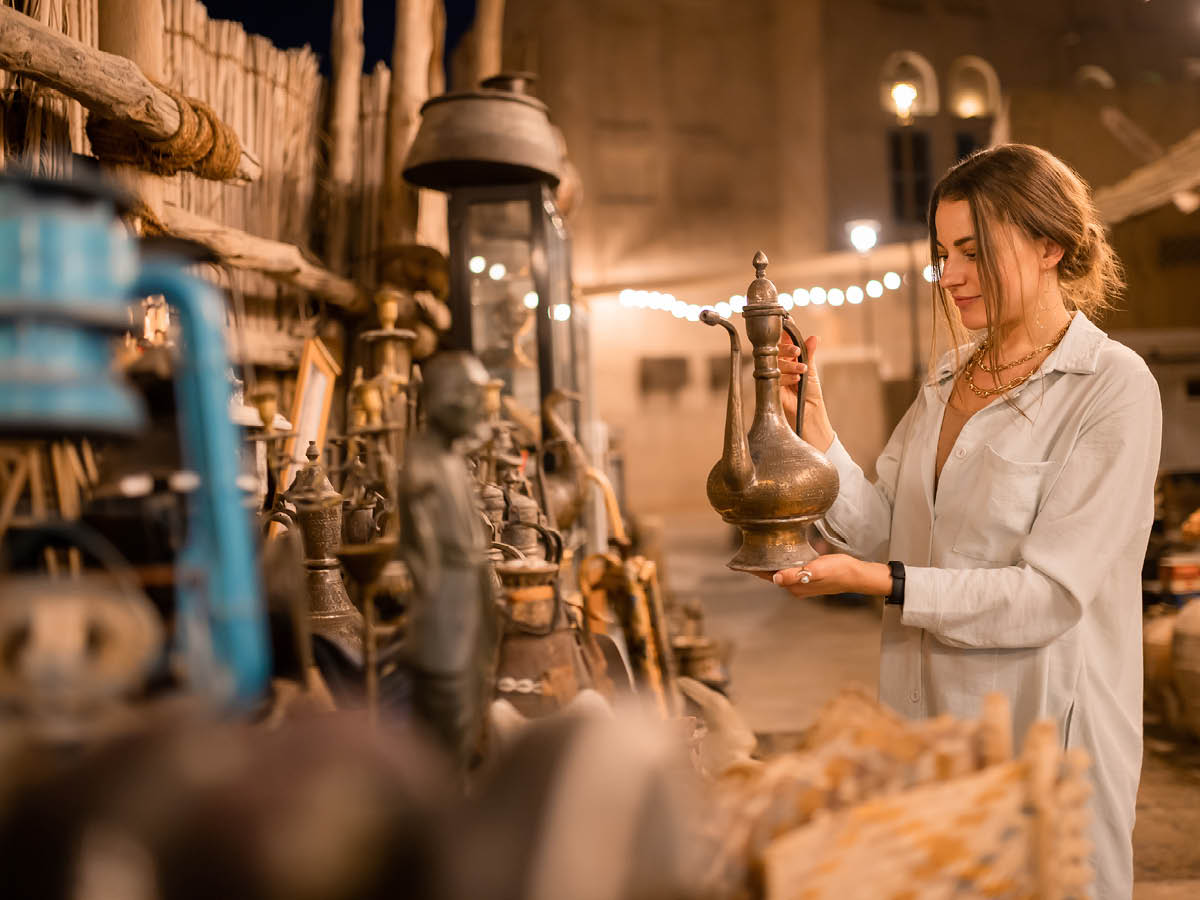 Eine Person hält einen antik wirkenden Metallkrug in einem Markt oder Geschäft, das mit verschiedenen Vintage-Gegenständen und Artefakten gefüllt ist. Im Hintergrund befinden sich Holzregale mit weiteren Metallobjekten wie Laternen, Statuen und Dekorationsstücken. Die Szene ist warm beleuchtet, mit sichtbaren Lichterketten im Hintergrund.