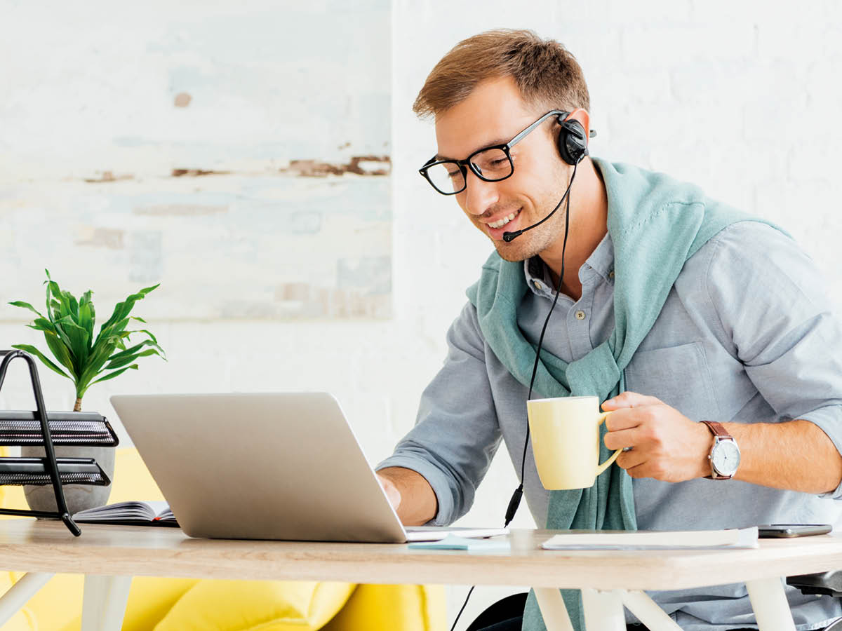 Eine Person sitzt an einem Schreibtisch und arbeitet an einem Laptop. Sie trägt Kopfhörer und hält eine gelbe Tasse in der Hand. Auf dem Schreibtisch stehen eine Pflanze sowie einige Bücher und Papiere.