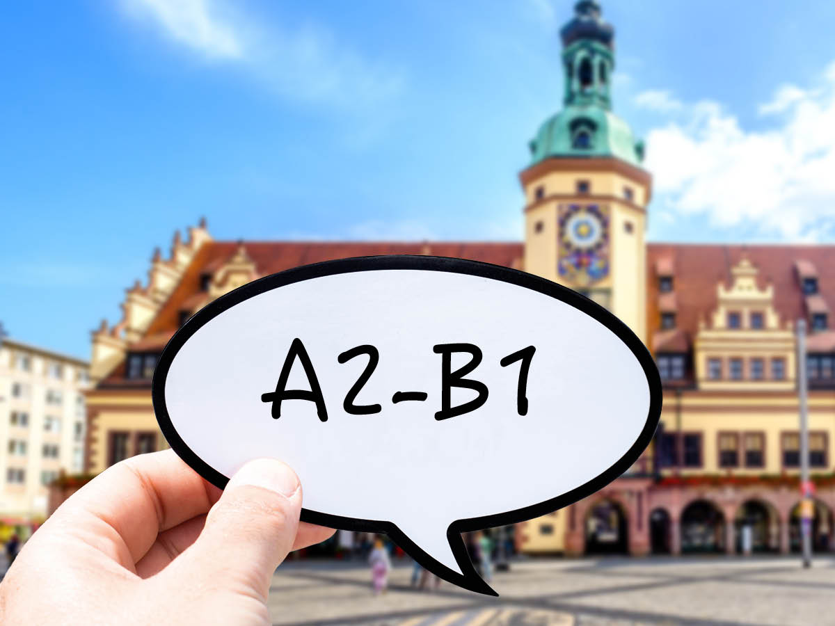 Eine Hand hält eine Sprechblase mit dem Text 'A2-B1'. Im Hintergrund ist ein historisches Gebäude mit einem Uhrturm und architektonischen Details zu sehen, wahrscheinlich in einem europäischen Stadtplatz.