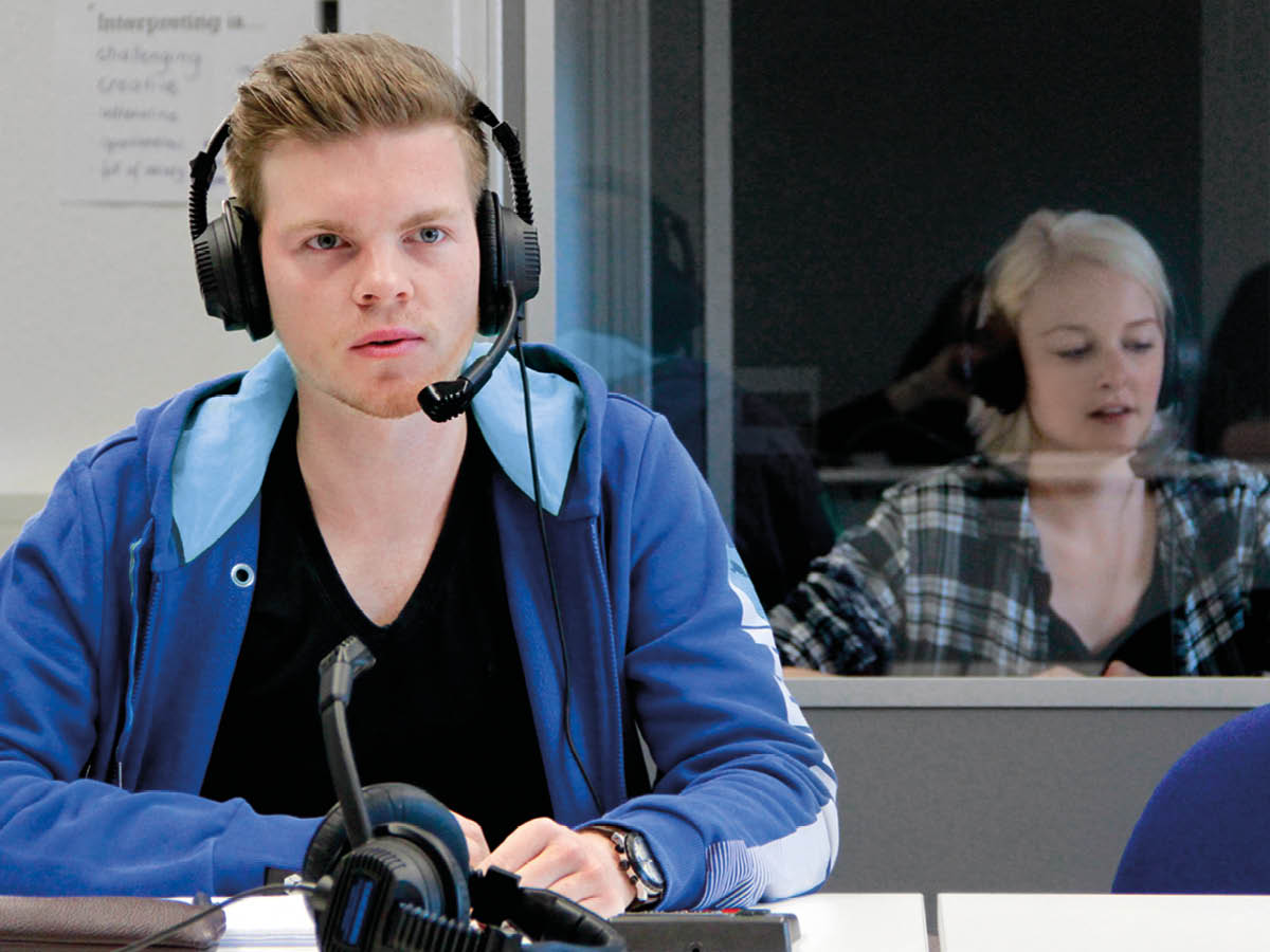 Zwei Personen befinden sich in einem Raum und tragen Headsets mit Mikrofonen. Die Person im Vordergrund sitzt an einem Schreibtisch mit einem Mikrofon und technischem Equipment. Die zweite Person sitzt hinter einer Glaswand im Hintergrund.