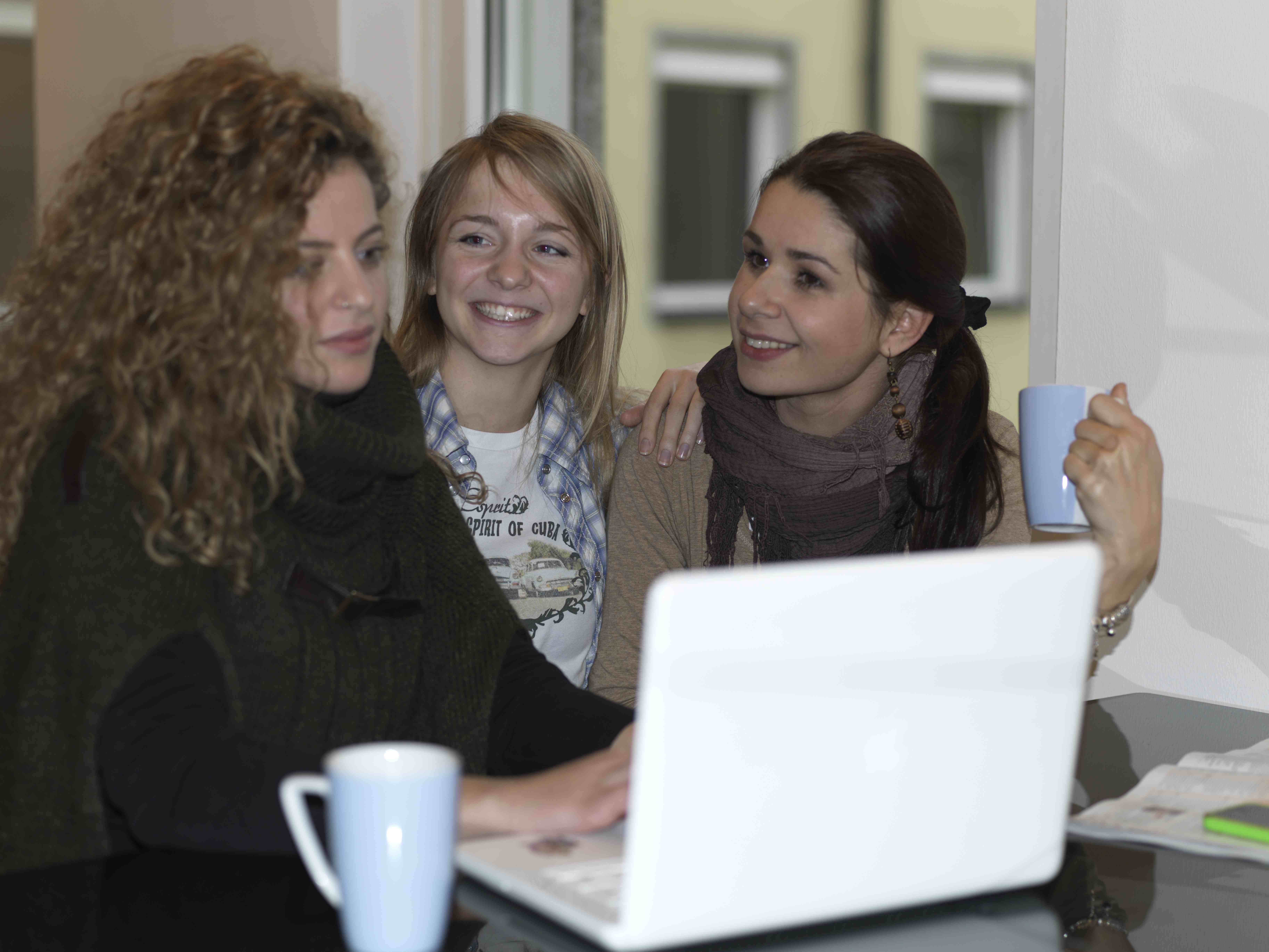 Fröhliche Zusammenarbeit am Laptop Drei Personen sitzen an einem Tisch mit Laptop und blauen Tassen