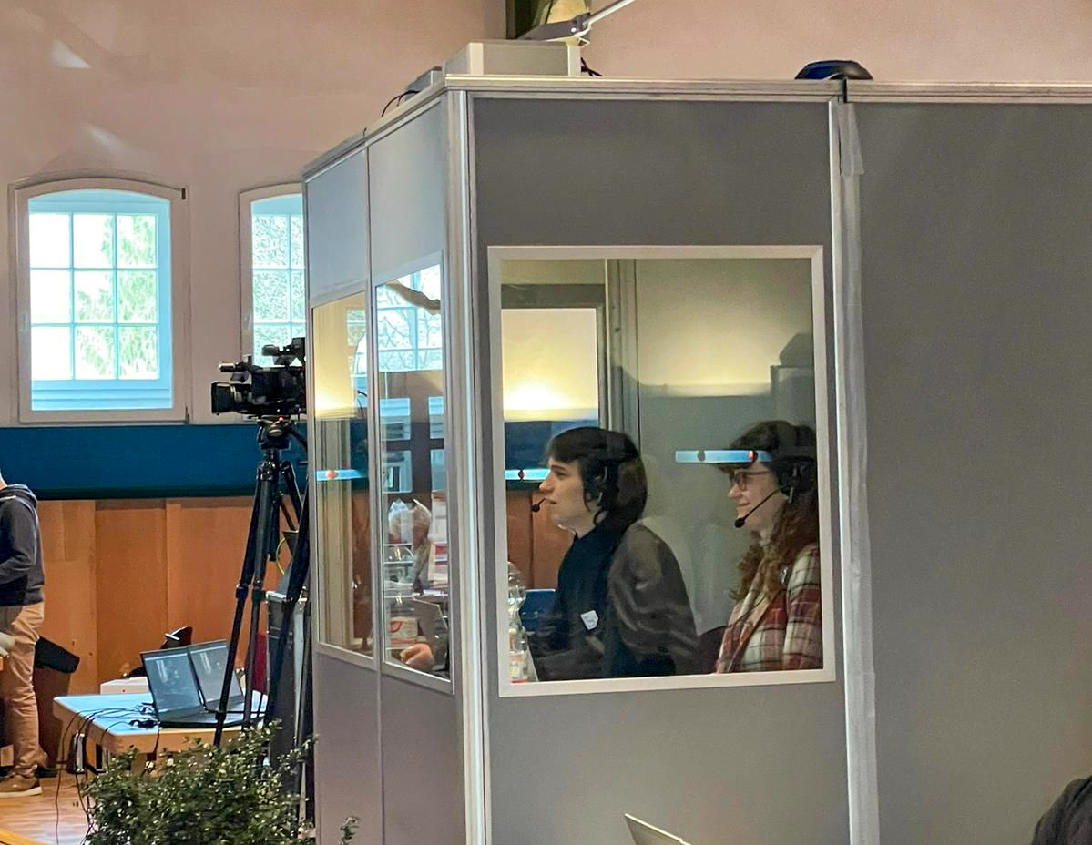 Zwei Personen mit Headsets in einer Dolmetscherkabine Zwei Personen mit Headsets in einer Dolmetscherkabine, umgeben von Kameraequipment in einem hellen Raum