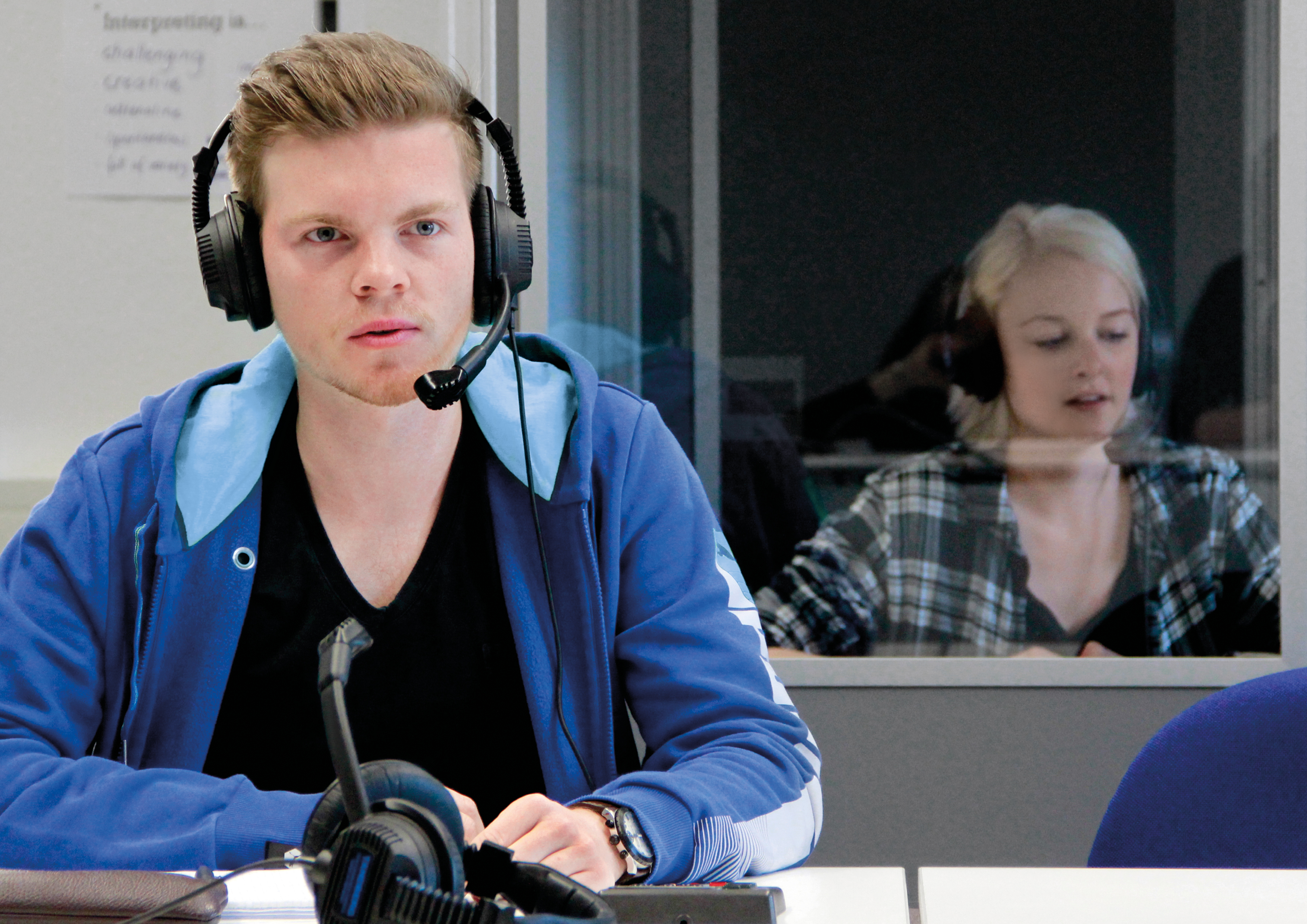 Dolmetscher bei der Arbeit Zwei Personen befinden sich in einem Raum und tragen Headsets mit Mikrofonen. Die Person im Vordergrund sitzt an einem Schreibtisch mit einem Mikrofon und technischem Equipment. Die zweite Person sitzt hinter einer Glaswand im Hintergrund.