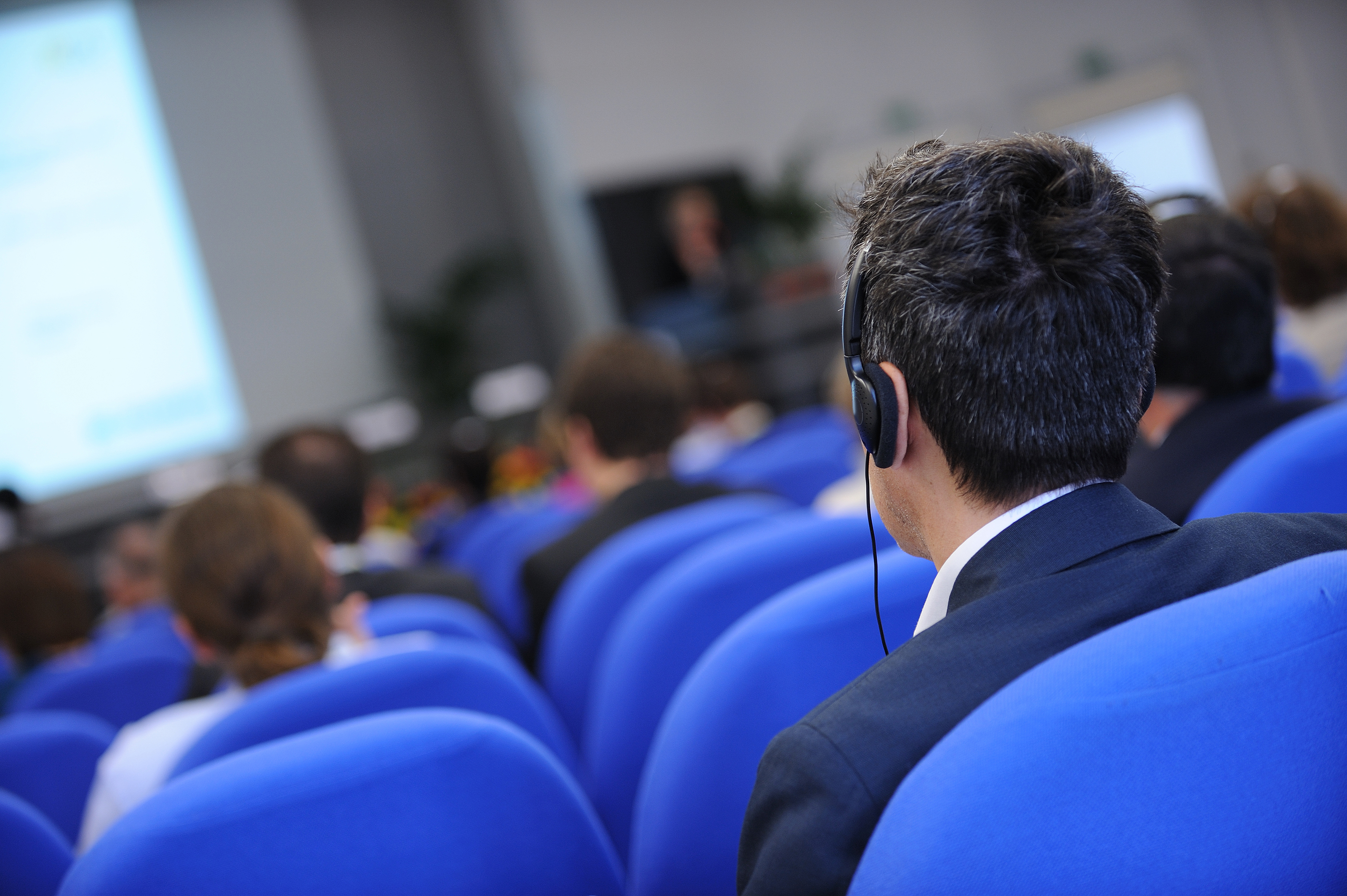 Teilnehmer sitzen auf blauen Stühlen bei einer Konferenz; im Vordergrund ein Mann mit Kopfhörern