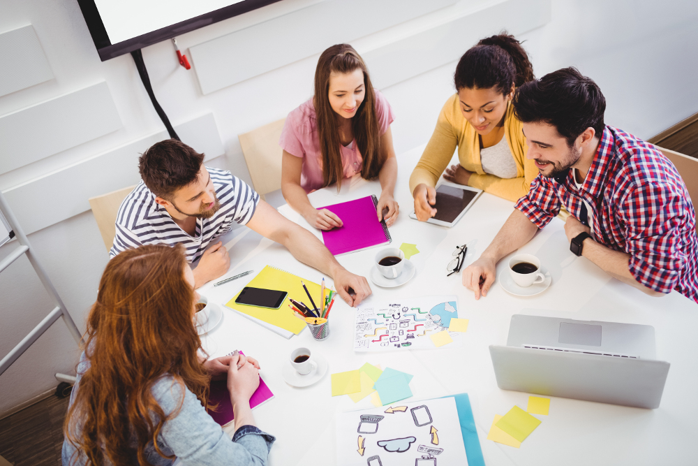 Personen sitzen um einen Tisch und führen eine Besprechung mit Notizbüchern, Tablets, Diagrammen und bunten Materialien.