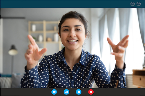 Im Web-Meeting Eine junge Frau in einer dunkelblauen Bluse mit weißen Punkten sitzt vor einem Bildschirm und gestikuliert mit den Armen. Sie scheint Teilnehmerin eines Web-Meetings zu sein. Im Hintergrund ist ein minimalistisch eingerichtetes Zimmer erkennbar.