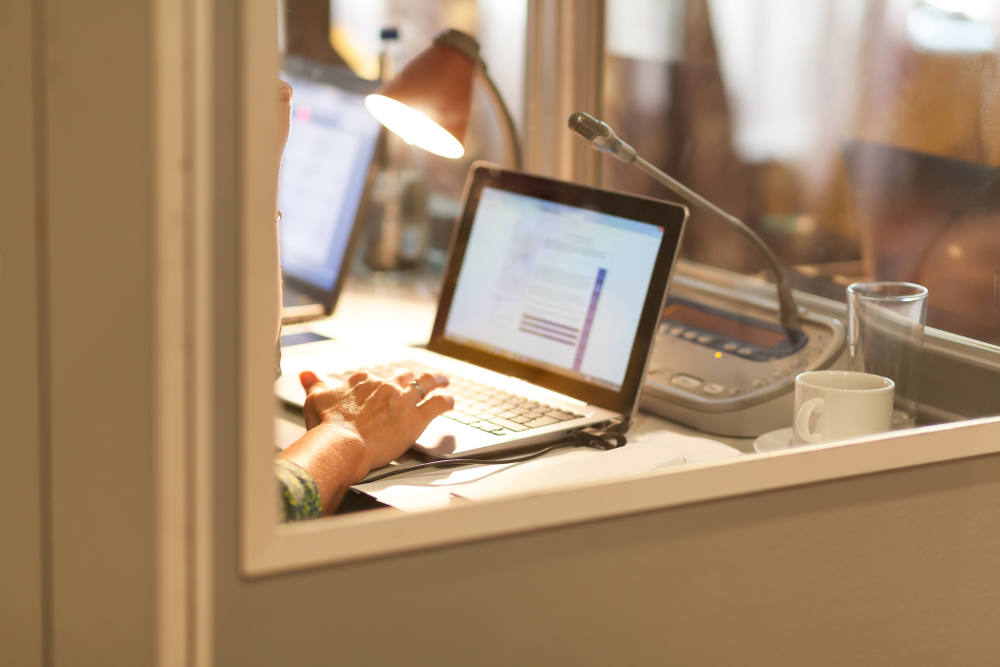 Person arbeitet an einem Laptop an einem Schreibtisch mit Mikrofon, Tasse, Glas und Papieren; eine Lampe beleuchtet den Bereich.